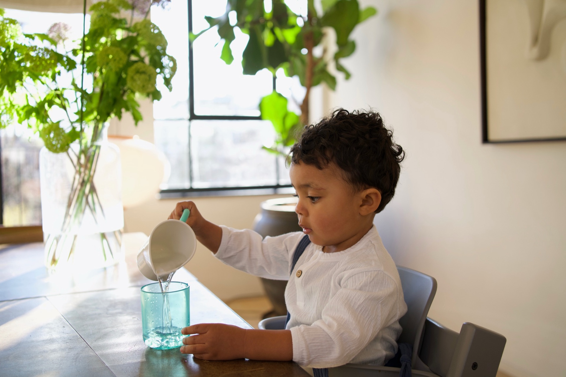 Peuter schenkt aan tafel voorzichtig water in een groen bekertje, met planten bij het raam